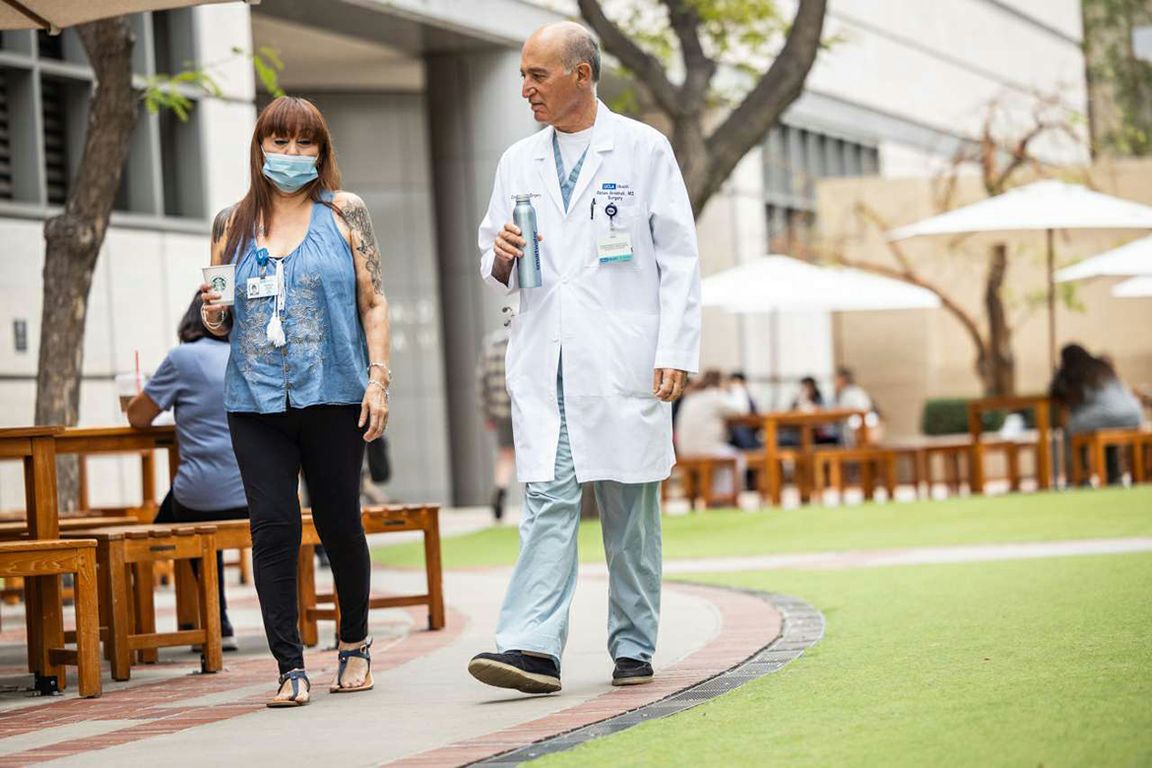 Photo: newsroom.ucla.edu .Dr. Abbas Ardehali, wearing a white lab coat over scrubs, walks outdoors with patient Patty Coles, who is wearing a