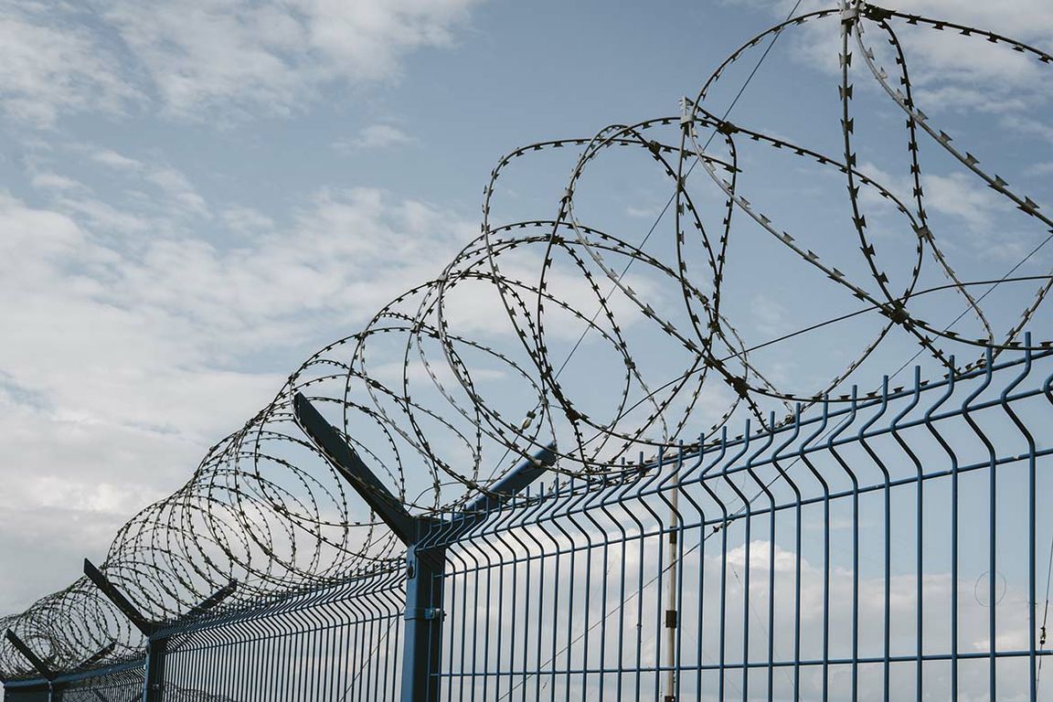Coils of razor wire atop a blue metal security fence against a partly cloudy sky.