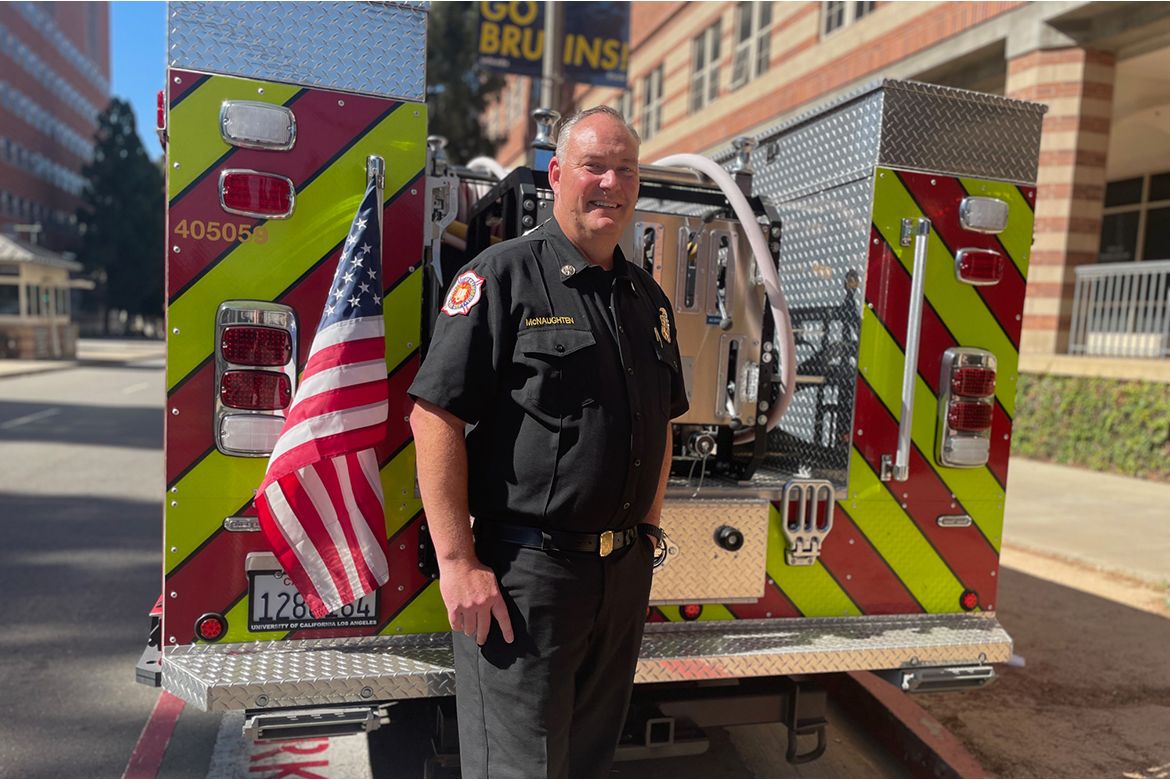 Steve McNaughten, in firefighter station wear uniform, stands smiling beside the rear of a fire engine displaying an American flag on a sunny city street.
