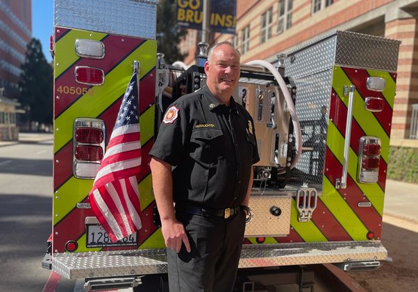 Steve McNaughten, in firefighter station wear uniform, stands smiling beside the rear of a fire engine displaying an American flag on a sunny city street.