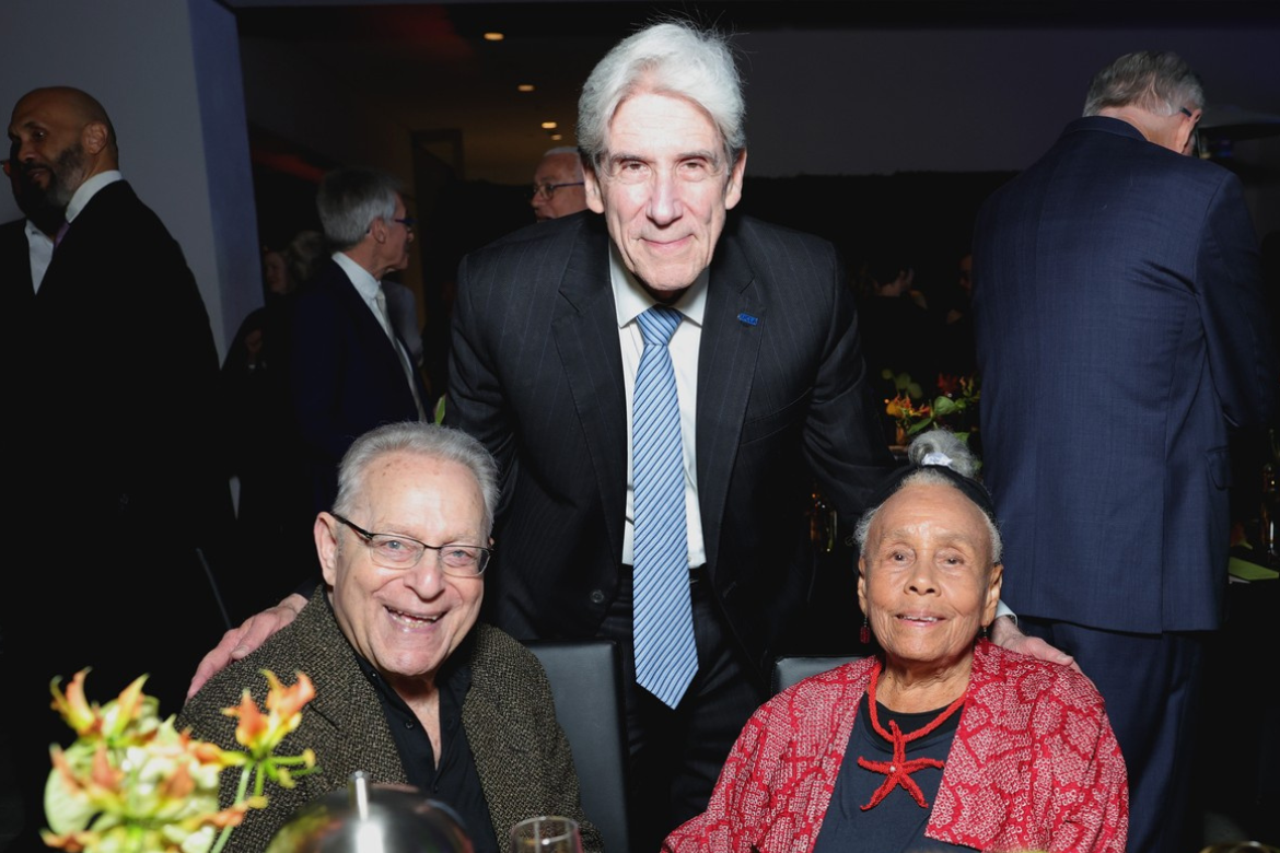 Joel Wachs and Betye Saar sit at a table with green and orange flowers as Chancellor Julio Frenk stands behind them
