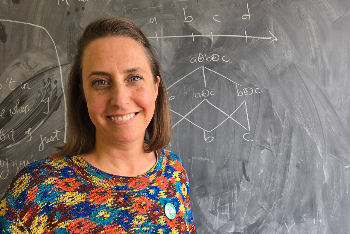 Close-up of Jessica Rett standing in front of a chalkboard covered with handwritten diagrams and symbols, facing the camera.