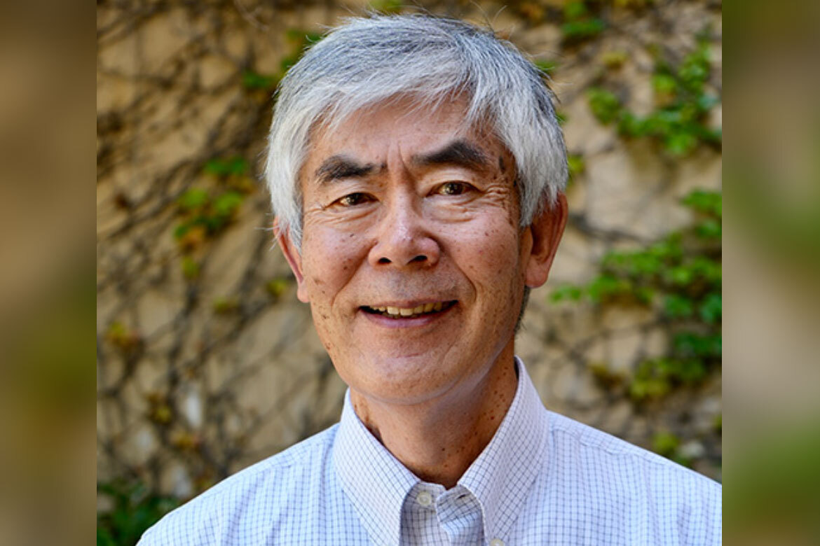 Portrait of Hiroshi Motomura wearing a light checkered button-down shirt, standing outdoors in front of a wall covered with green vines.
