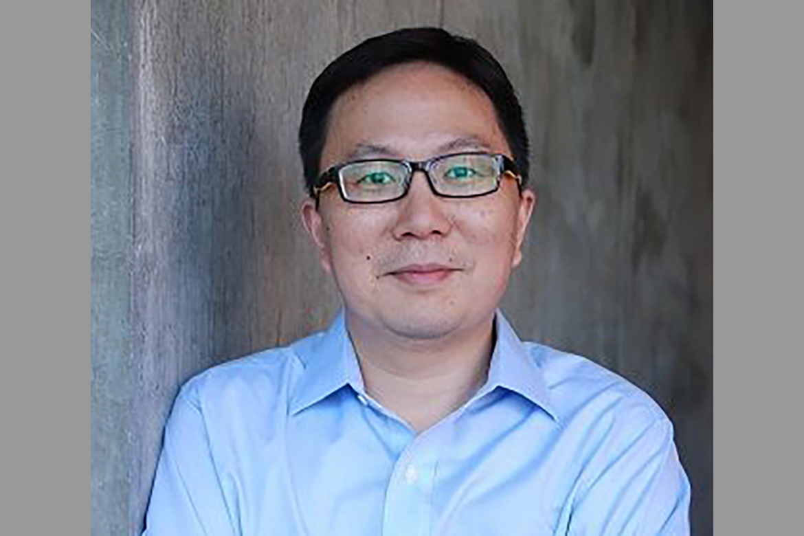 Portrait of Xiangfeng Duan wearing black-framed glasses and a light blue button-down shirt, facing the camera with a slight smile against a softly blurred gray background.