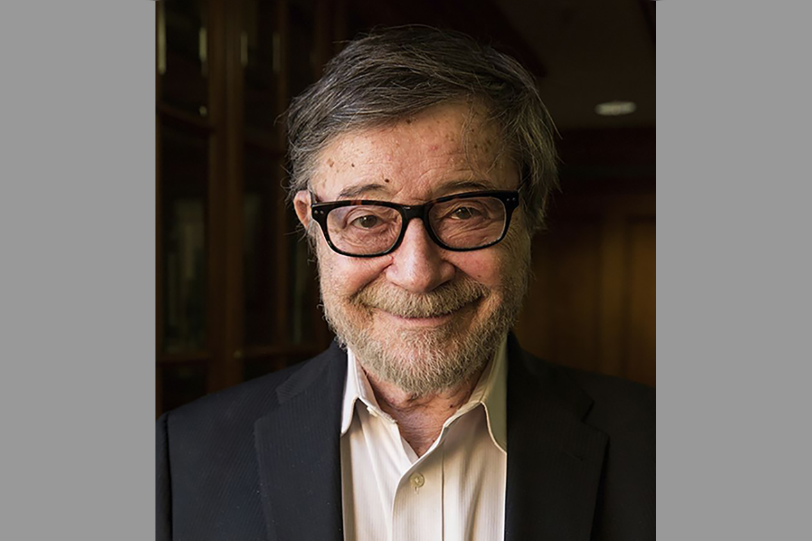 Portrait of Judea Pearl, wearing a dark blazer, light shirt and black-rimmed glasses, indoors against a softly blurred wood-paneled background.