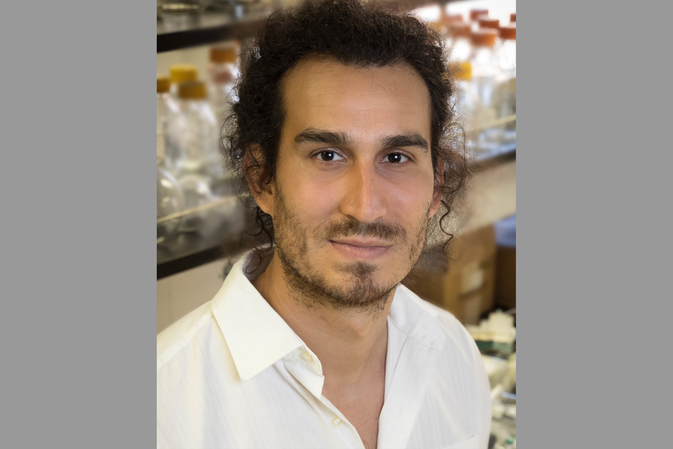Portrait of Matthew Nava in pale yellow button-down shirt standing in front of glass bottles in the lab.