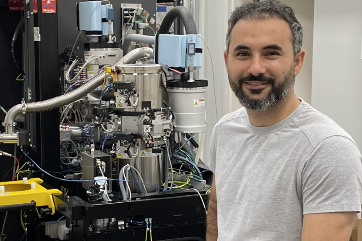 Navid Bavi, in gray T-shirt, sits next to a large scientific instrument with stainless steel chambers, hoses and control modules in a laboratory, facing the camera.