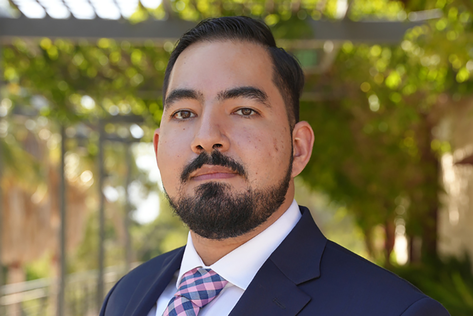 Portrait of Steven Chavez in a navy suit, white shirt and pink-and-blue tie, standing outdoors with trees and a pergola blurred in the background.