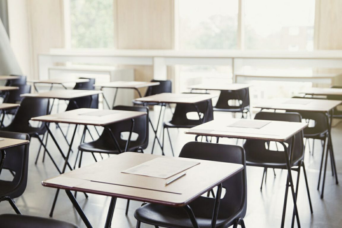 An empty classroom with rows of student desks and chairs illuminated by daylight from large windows.