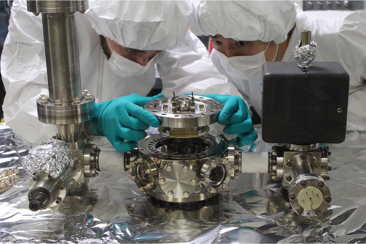 Michael Bareian, left, and Yiyang Zhi in cleanroom suits and gloves carefully install a circular scientific instrument onto a stainless-steel vacuum chamber surrounded by precision laboratory equipment.
