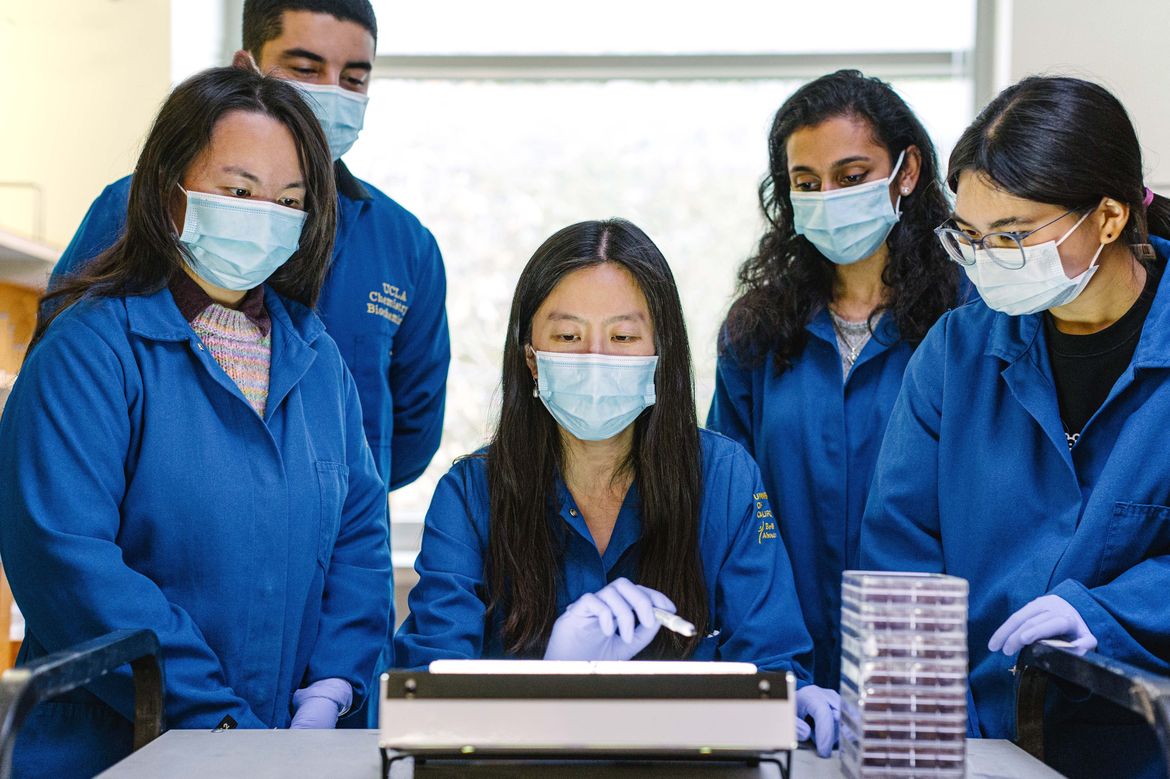 Melody Li, centered, gestures to a benchtop instrument while four masked researchers in blue lab coats watch closely. A stack of clear culture plates sits on the lab bench beside them. 