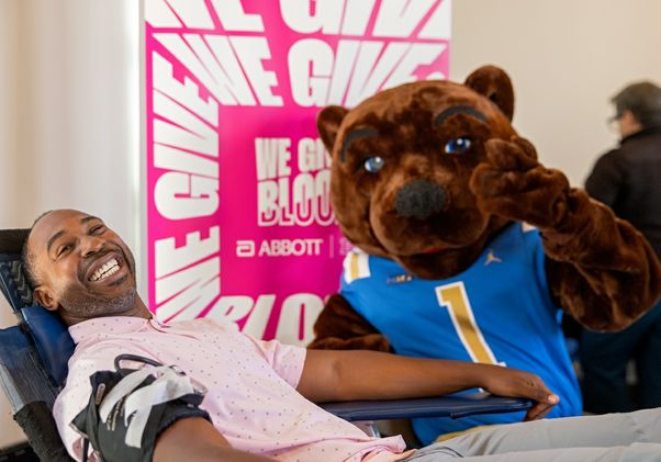 UCLA Athletic Director Martin Jarmond reclines in a blood-donation chair with a bandage on his arm as Joe Bruin, wearing a UCLA jersey, stands beside him and gives a thumbs-up.