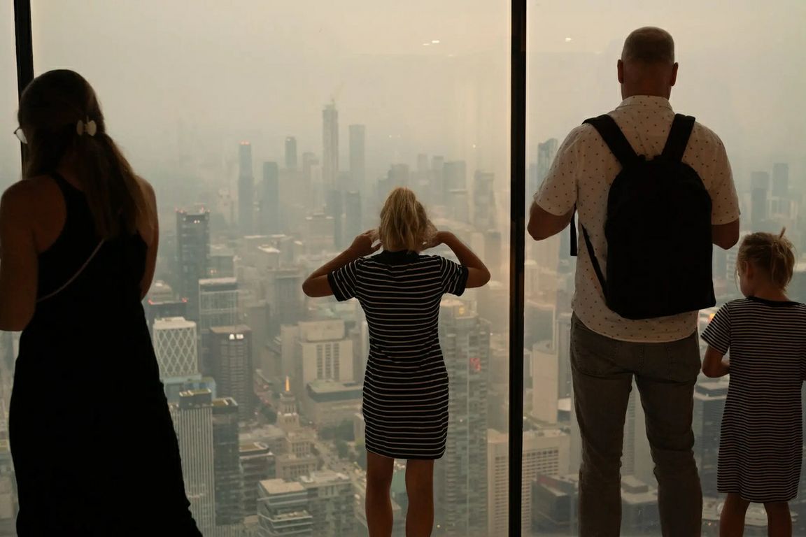 People look through floor-to-ceiling windows at a city skyline obscured by wildfire smoke and haze.