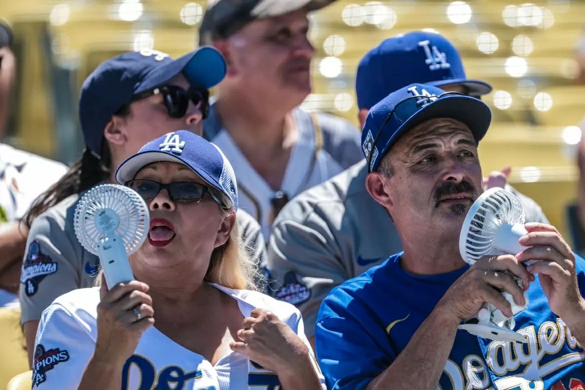 A group of Los Angeles Dodgers fans in team apparel use handheld fans to cool themselves in the stands.