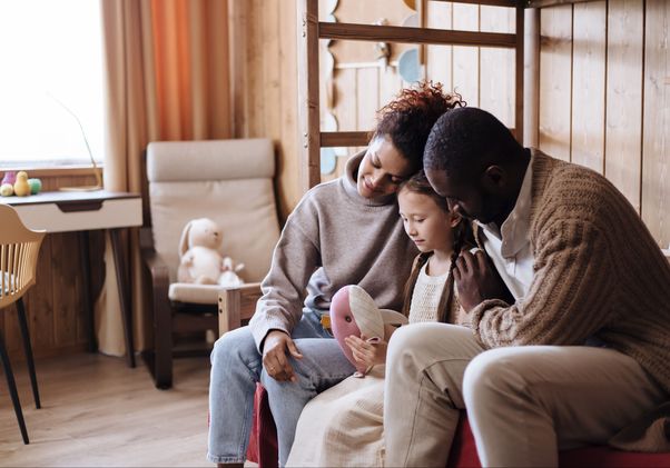 A family sits closely together on a sofa, reading a picture book with a young girl in a cozy wood-paneled room.