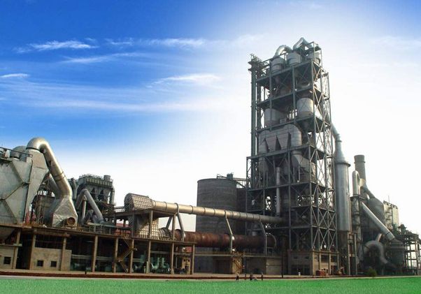 Industrial cement production facility with a rotary kiln, clinker-processing equipment, large ducts, towers and interconnected machinery beneath a blue sky.