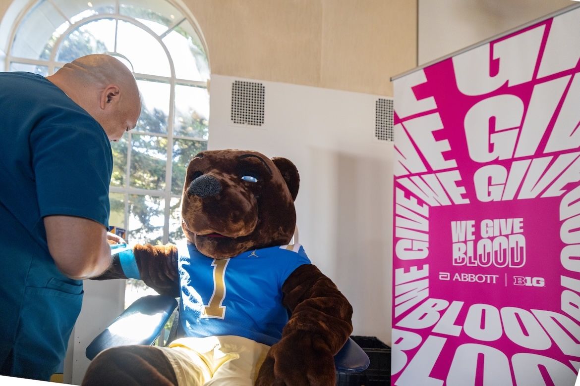 Joe Bruin sits in a blood donation chair as a staff member prepares his arm, with a bright pink “We Give Blood” campaign banner beside him.