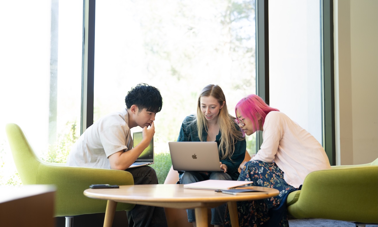 Alt text: Three UCLA students gather around a laptop, working together in a bright campus study space.