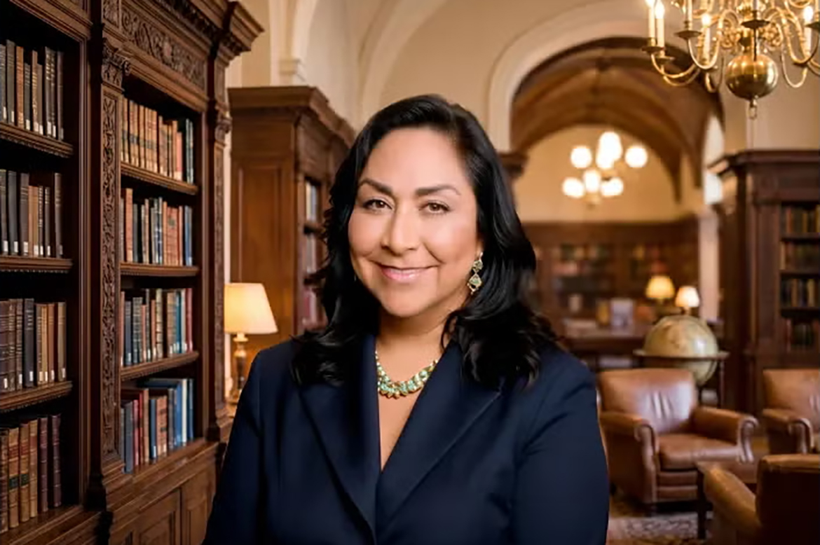 Marta Segura wearing a navy blazer in a library, surrounded by ornate wooden bookshelves, leather chairs, warm lighting and a globe.