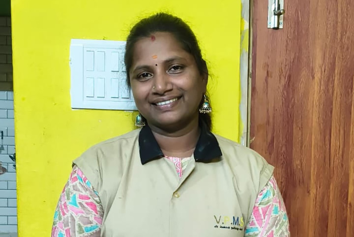 Mohana Sundari smiles at the camera, wearing a beige vest over a colorful patterned shirt, with a bright yellow wall and wooden door behind her.