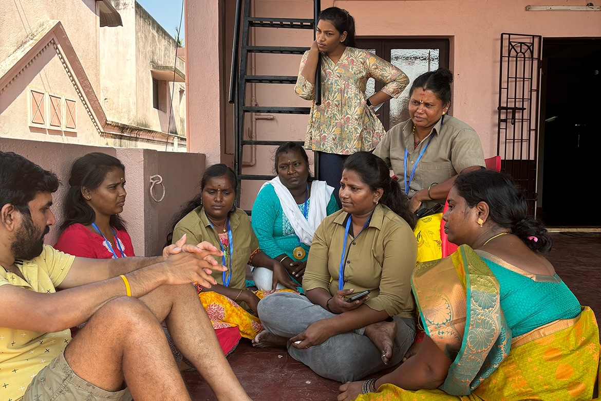 Mohana Sundari, seated with several women outside a building, speaks with a man as the group listens.