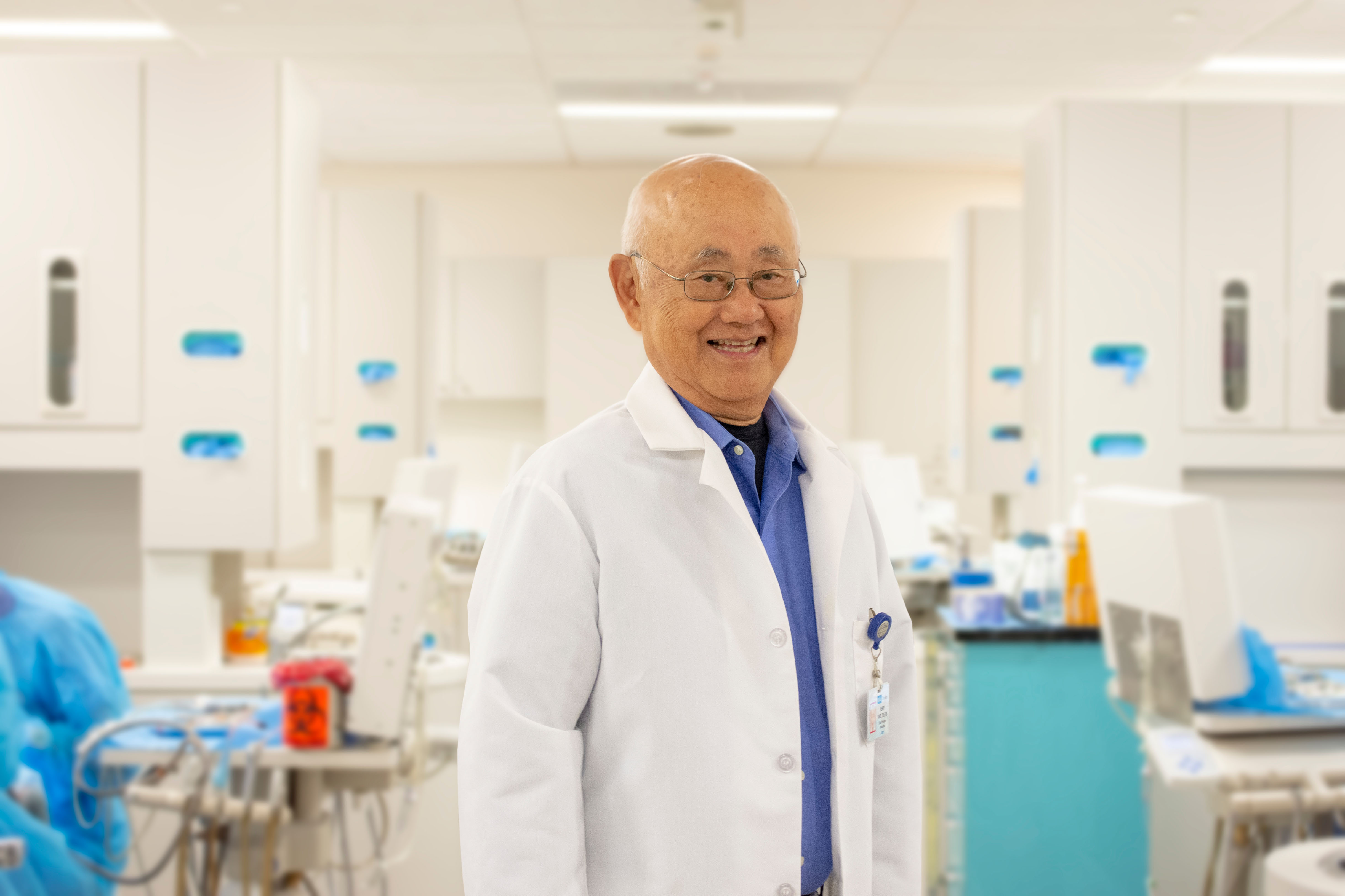 Henry Takei wears a white lab coat and glasses while standing in a bright laboratory with medical equipment and workstations visible in the background.