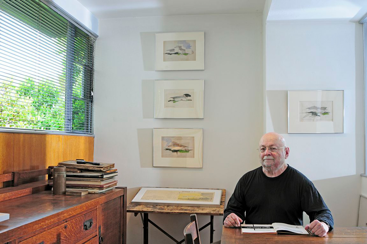 Architectural historian Thomas Hines sits at a table with an open book in his Richard Neutra–designed apartment, surrounded by framed artwork, wooden furnishings, stacked books and sunlight filtering through window blinds.