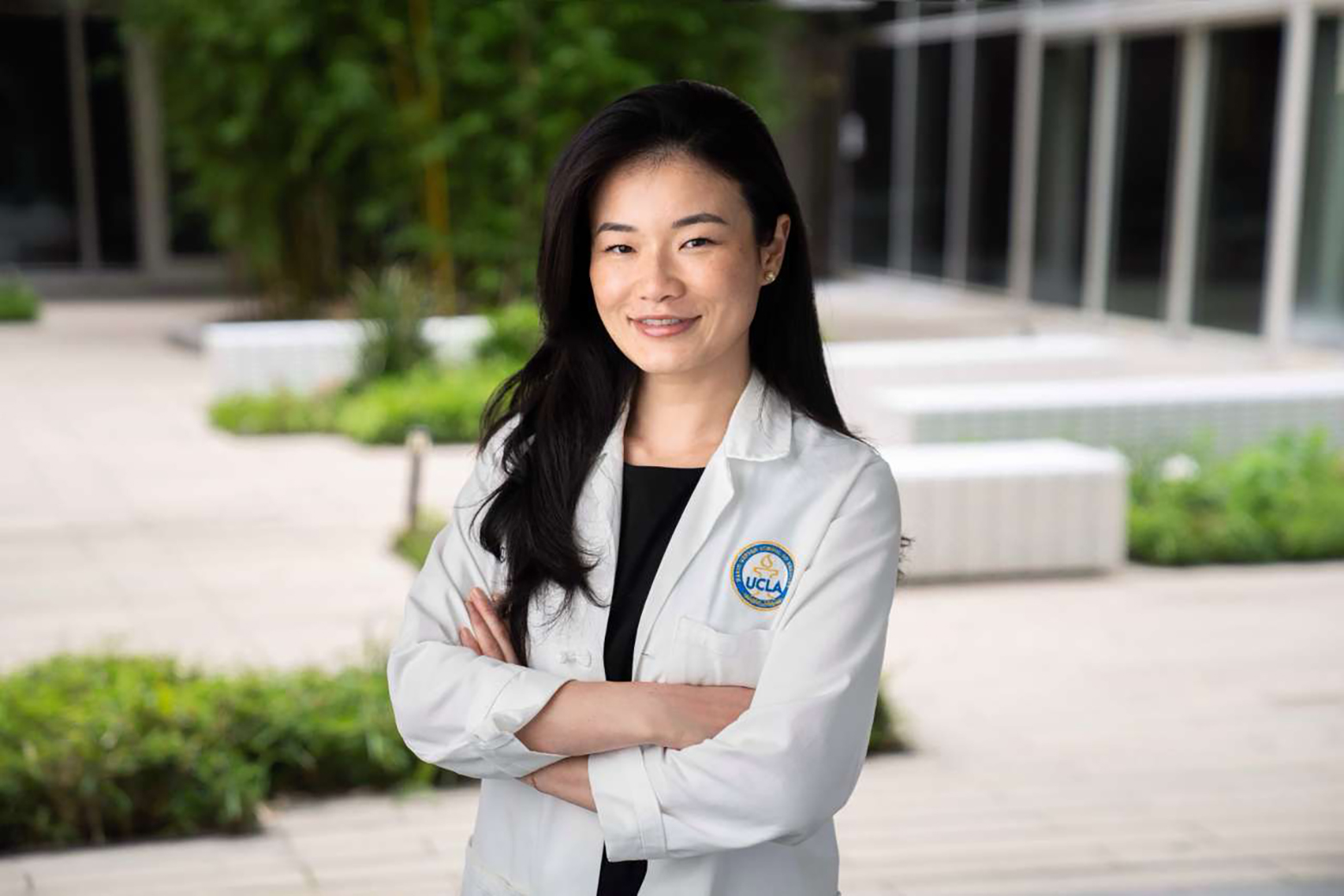 Jie Deng, in a white lab coat, stands outdoors with her arms crossed, smiling in front of a modern campus building.