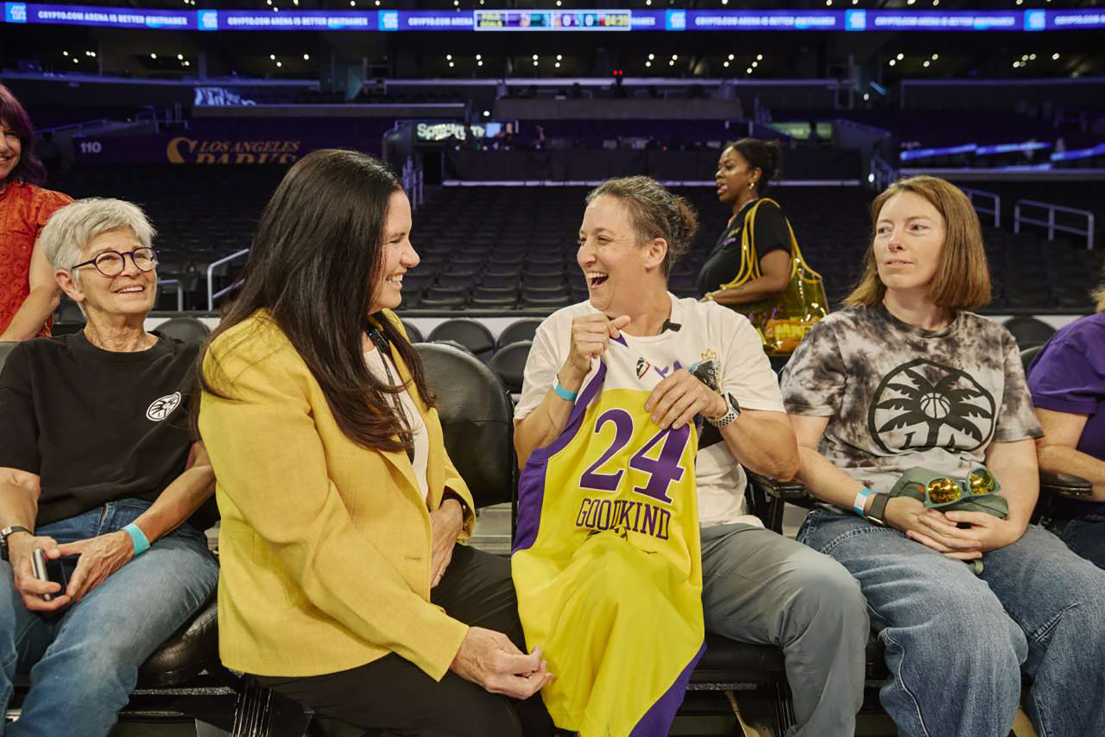 From left: Barbara Frankin in black T-shirt and jeans, Sharon Hame in yellow jacket, Laura Goodkind holding a Sparks jersey and Jennie Coyne in graphic T-shirt sit courtside at a basketball arena.