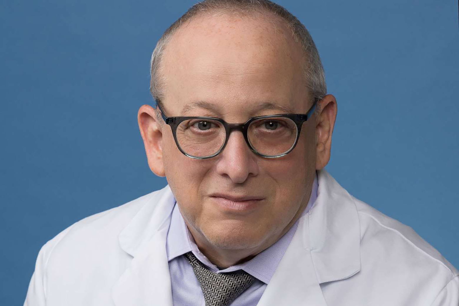 Neil Silverman wearing glasses, a white lab coat, dress shirt and patterned tie, facing the camera against a solid blue background.