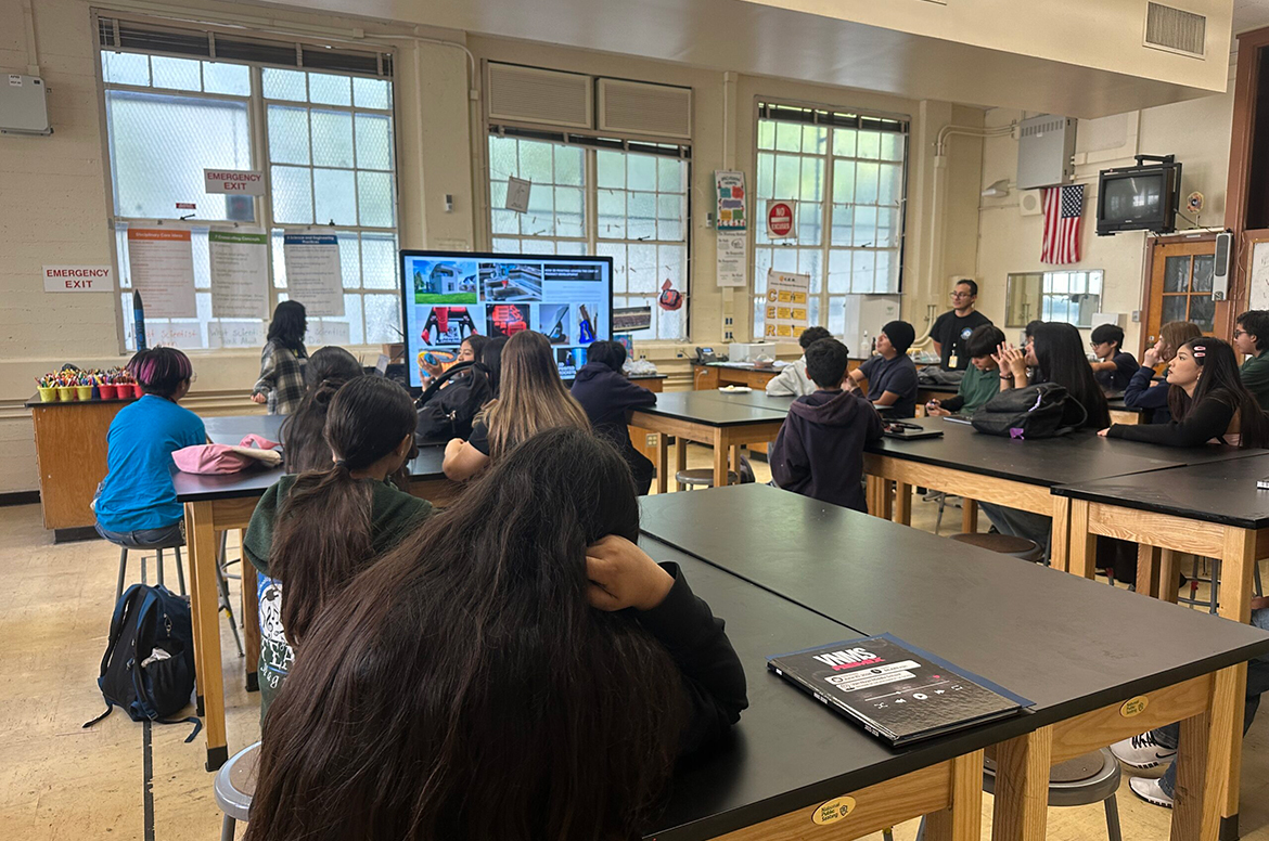 Students sit at tables in a classroom while a teacher presents images on a monitor during a group lesson.