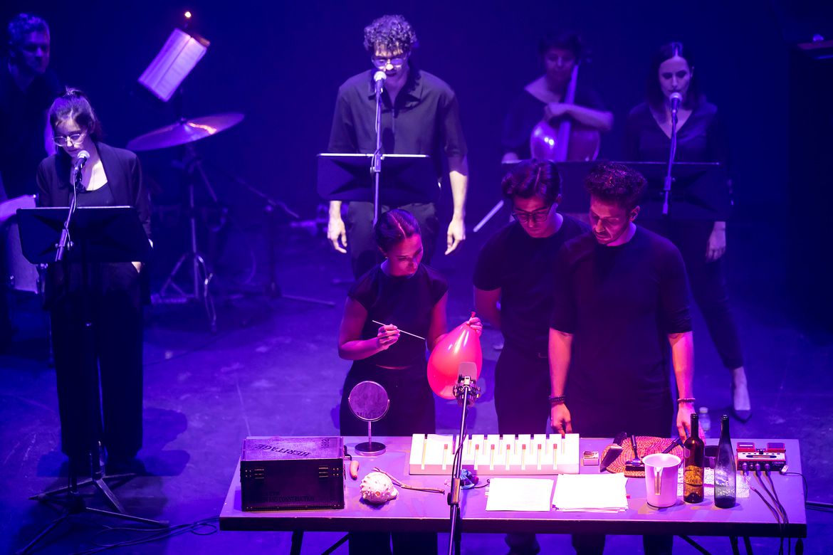 Musicians perform onstage under purple lighting, with singers at microphones and performers gathered around a table of instruments and props.