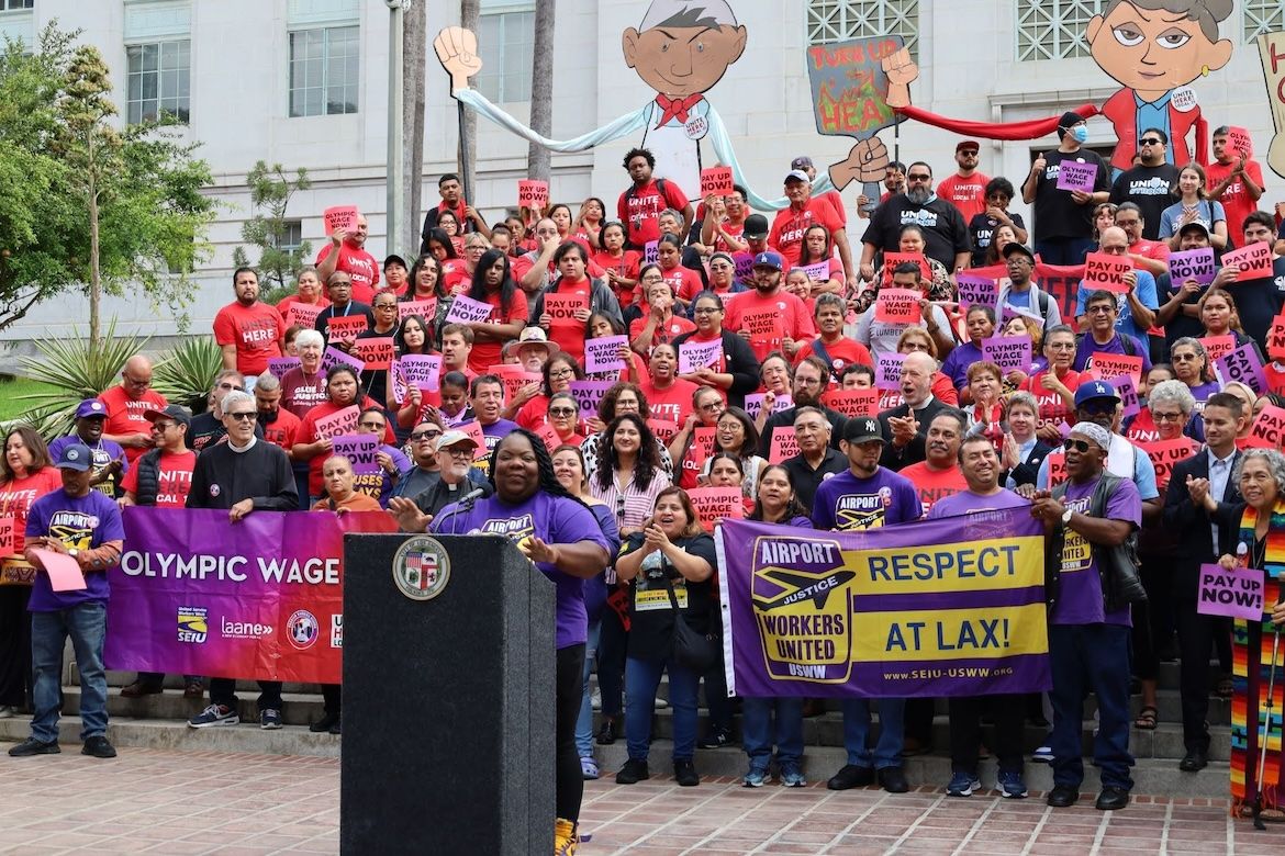 A large crowd of union workers and supporters gathers on the steps of Los Angeles City Hall, holding signs calling for higher wages and respect for workers as a speaker addresses the group from a podium.