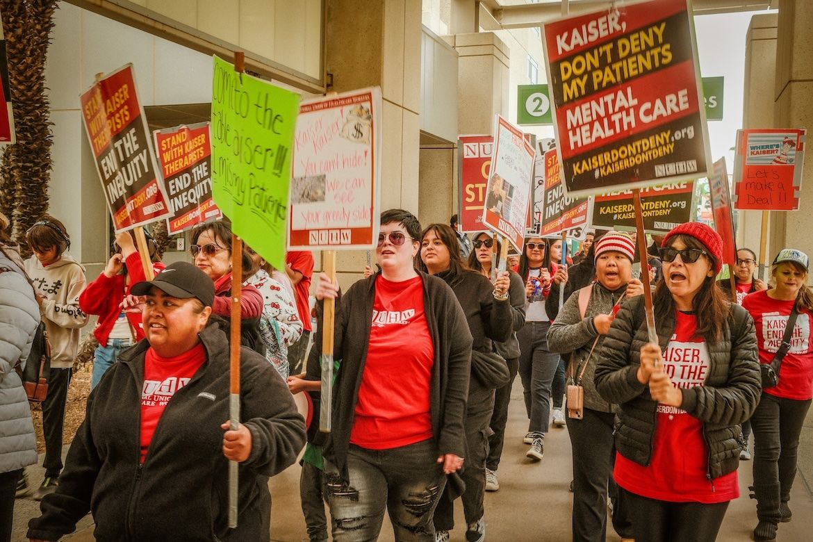 A group of Kaiser health workers marches with protest signs calling for an end to inequities in care and urging the health system not to deny patients mental health services.