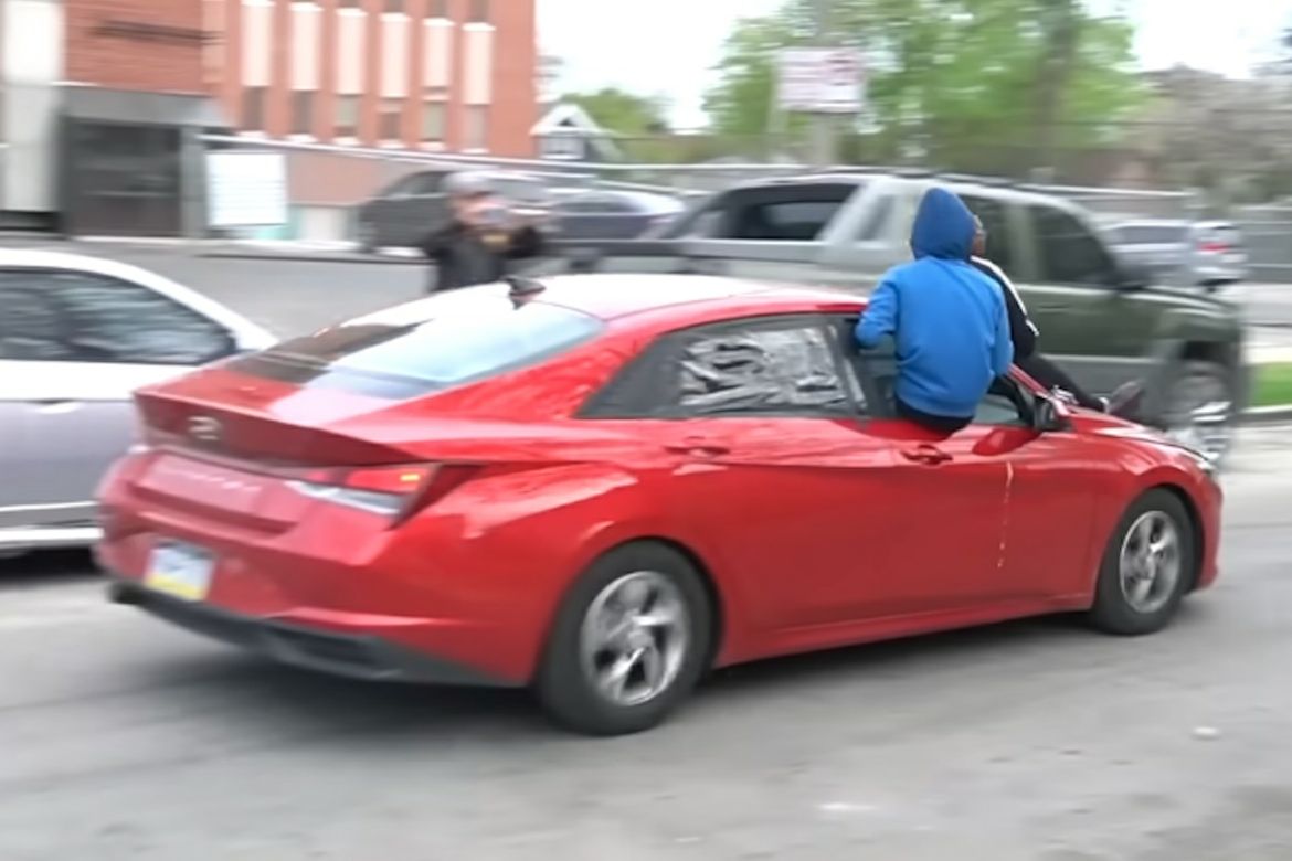 YouTube video still of a person in a blue hoodie riding partly outside a red car as so-called “Kia Boys” joyride in a stolen vehicle in 2022.
