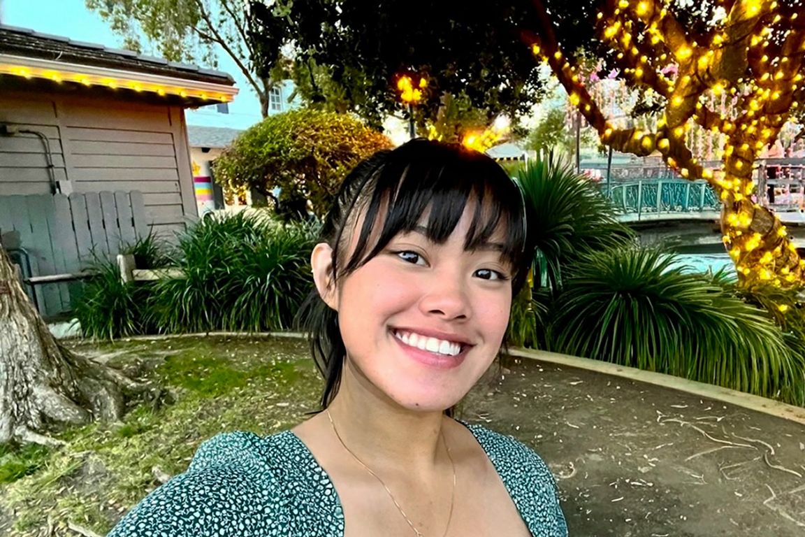UCLA student Deanna Placa smiles for a selfie outdoors, surrounded by trees and greenery decorated with string lights.