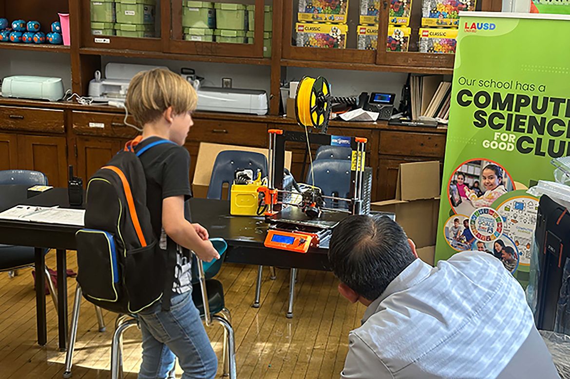 Student examines a 3D printer in a classroom while an adult observes; educational materials and a computer science club banner are visible.