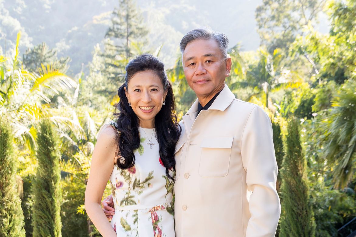 From left: Shirley Wang, in a floral dress, and Walter Wang, in a cream-colored jacket, pose outdoors in a sunlit garden, surrounded by lush greenery. 