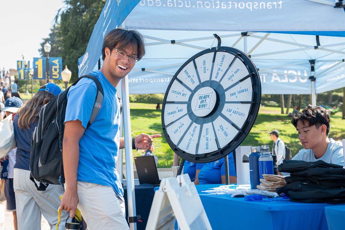 A smiling student in a blue T-shirt points toward a prize wheel at a university event booth, with other students and staff nearby.