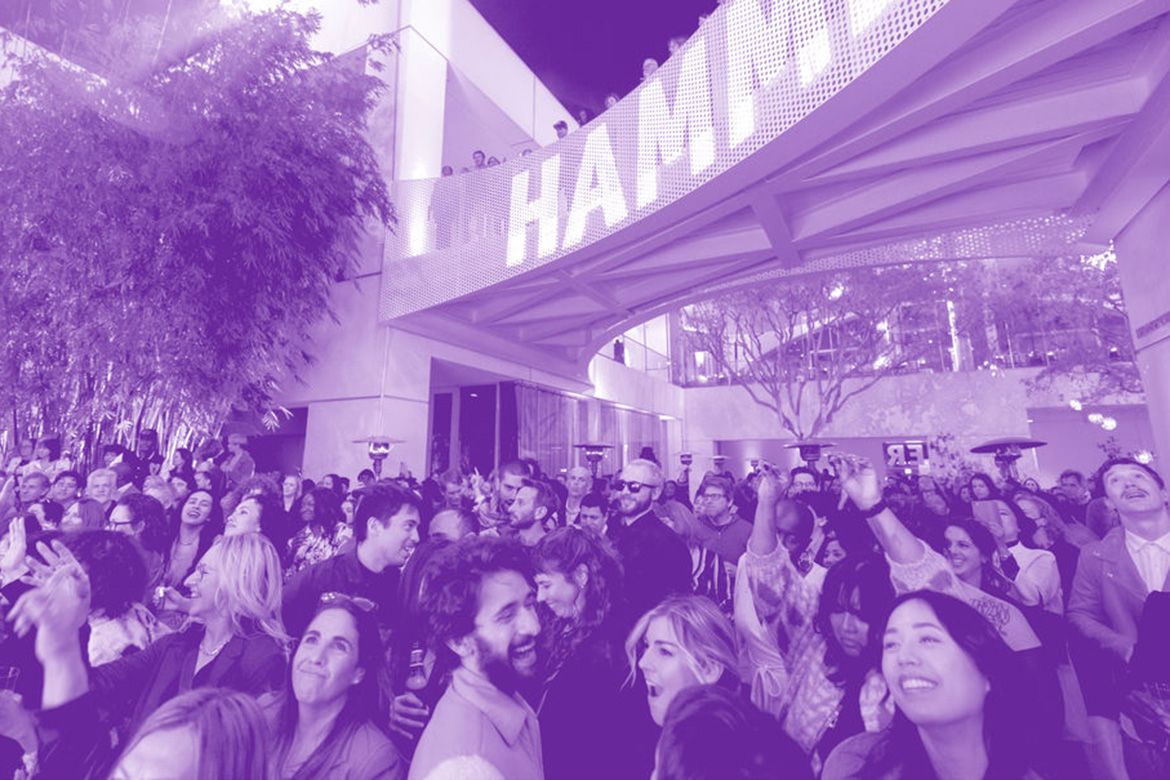 Crowd of people gathered outdoors beneath a modern building, with trees, cameras, and a large illuminated sign overhead, all tinted purple.