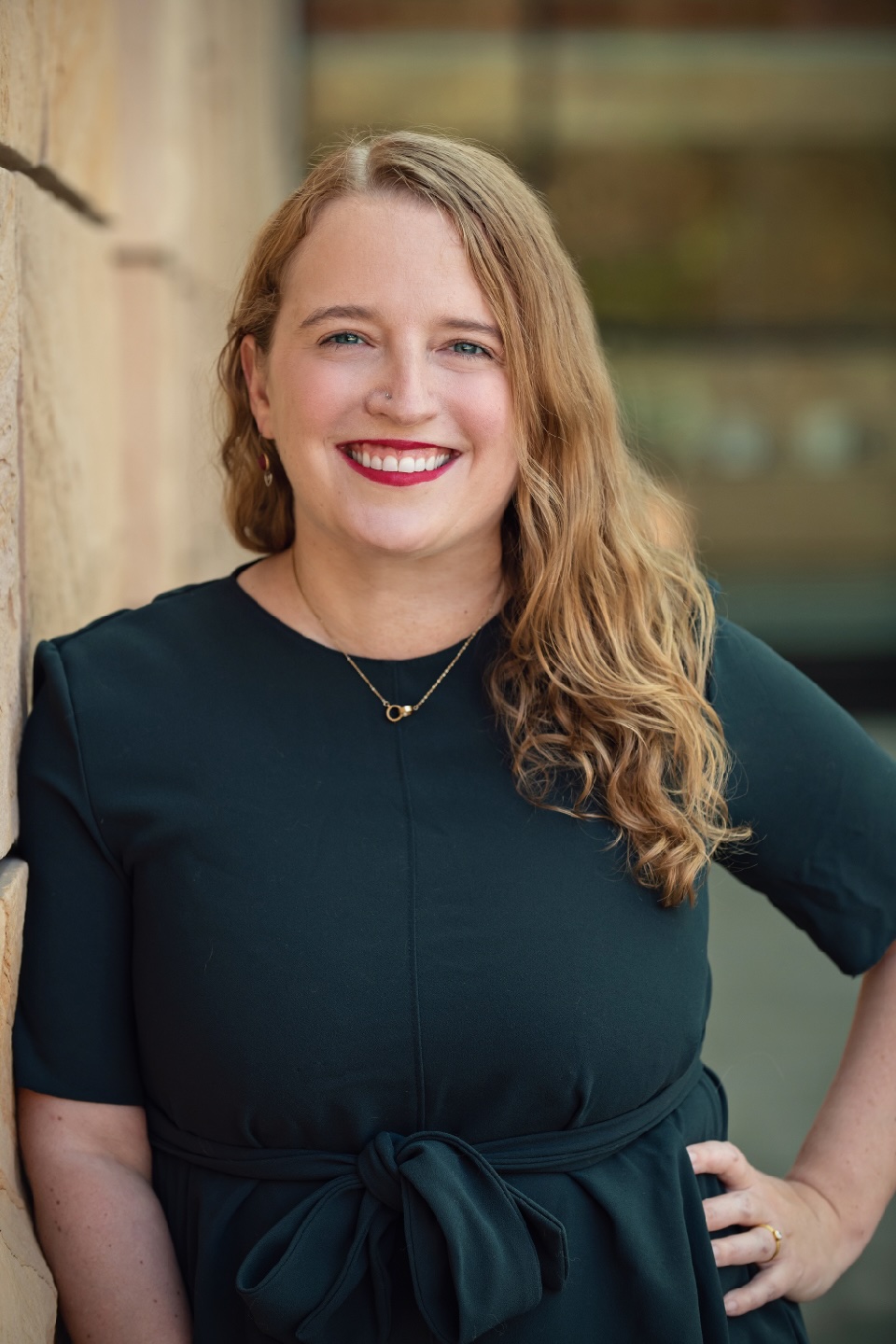 Portrait of UCLA's Catherine Bonesho, smiling woman with long, wavy blond hair, wearing a dark green top and delicate necklace and leaning against a stone wall.