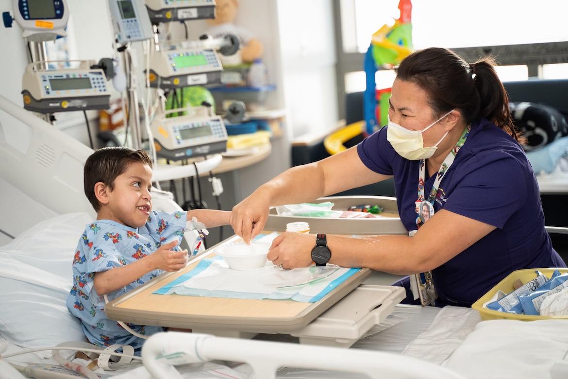 A health care worker assists a young patient sitting in a hospital bed, with medical equipment visible nearby.