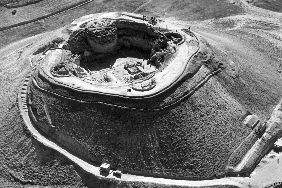 Black-and-white aerial view of Herodium, an ancient palace-fortress built atop a steep, rounded hill in the Judean Desert, showing its circular fortifications, excavated ruins and surrounding arid landscape.