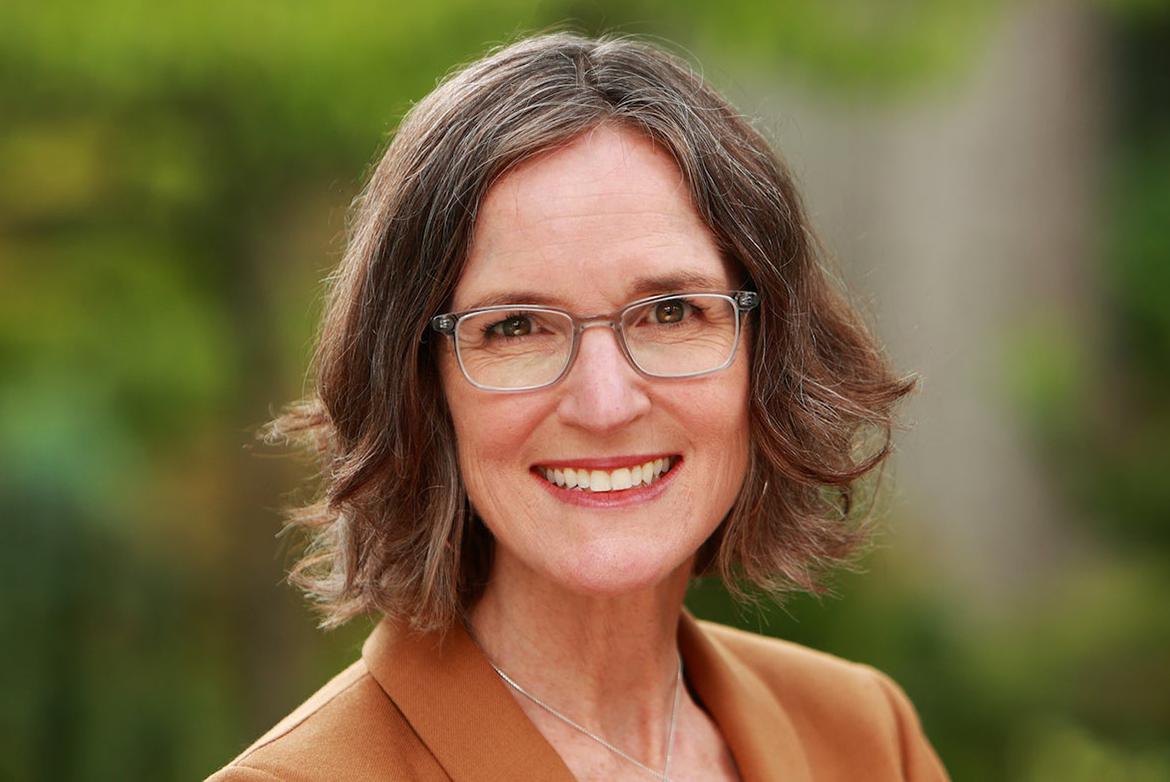 Close-up of Megan Mullin, wearing glasses and a brown jacket, photographed outdoors against a softly blurred green background.