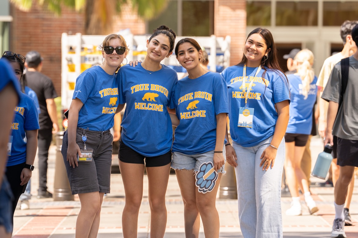 Four UCLA students wearing blue “True Bruin Welcome” T-shirts pose together outdoors on campus during a welcome event.