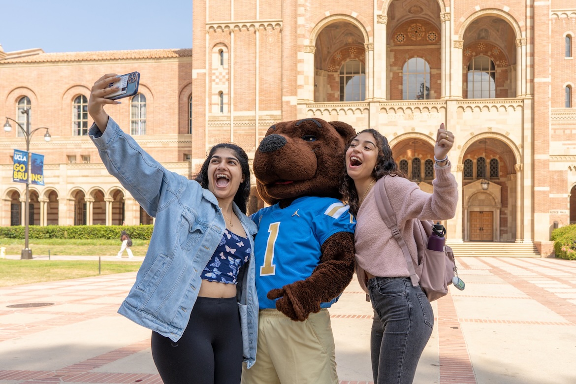 Two UCLA students take a selfie with the Joe Bruin mascot in front of Royce Hall.