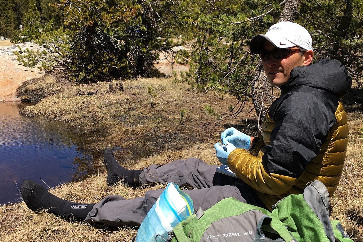 Dave Daversa wears blue gloves as he sits beside a pond in a forested area, handling a small object near outdoor gear.