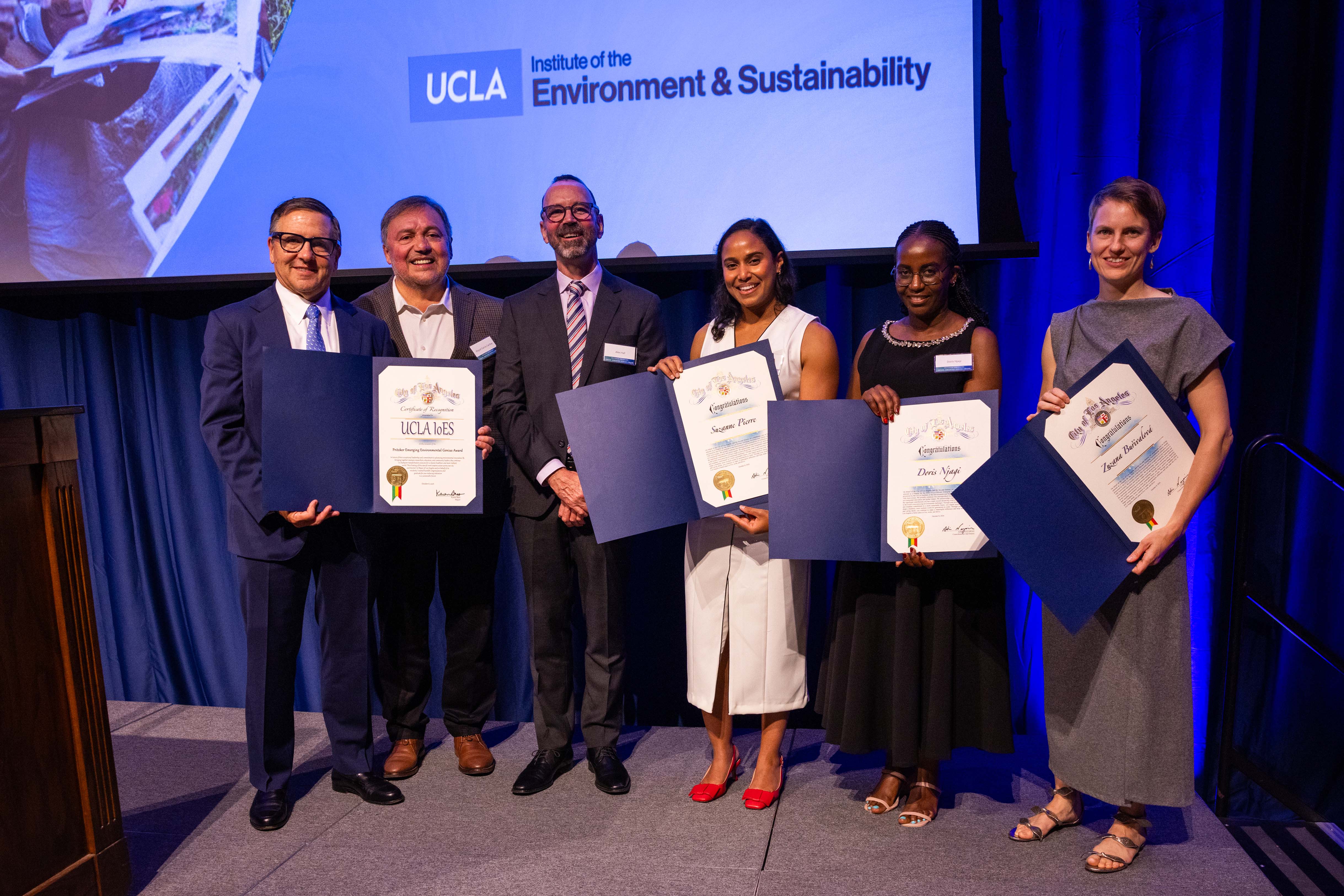 Six people pose onstage, some holding certificates in front of a UCLA Institute of the Environment & Sustainability backdrop, with blue stage lighting and curtains.