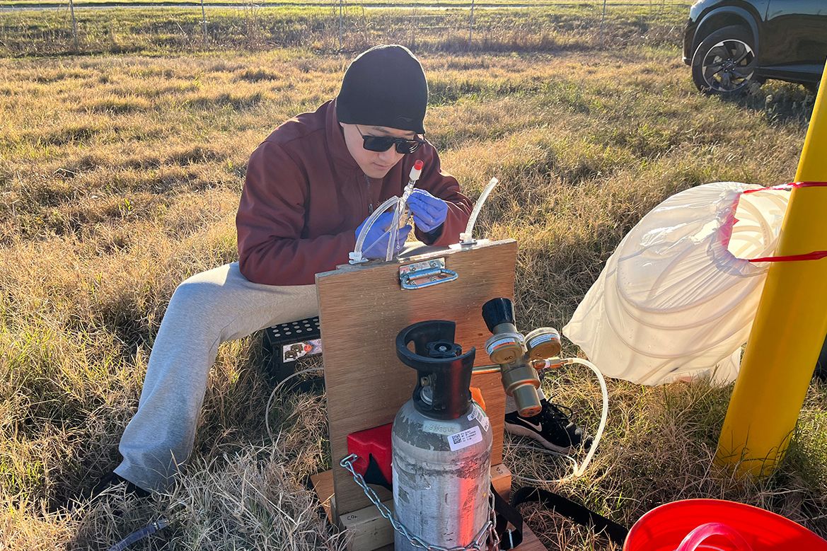 Jiarui Liu, in protective clothing, works with equipment and gas cylinders beside a large yellow structure in a grassy field.