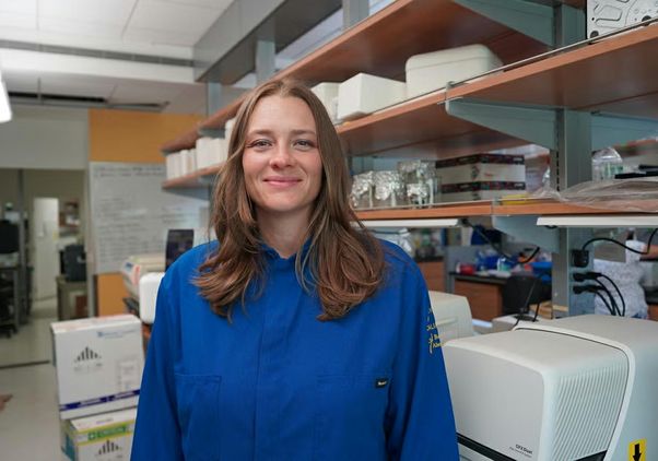 Tara TeSlaa, in a blue lab coat, stands smiling in a laboratory, surrounded by shelves of equipment, supplies and scientific instruments.